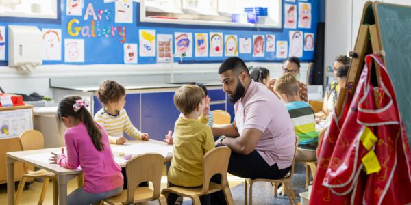 Pre-school students sitting in an art classroom being taught by a teacher. The classroom is colourful and the children are sat at a big table.