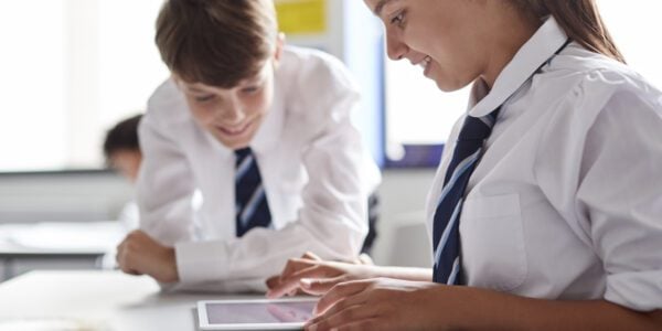 Two schoolchildren in white shirts and striped ties are sitting at a desk in a classroom, engaged with a tablet. The girl in the foreground is smiling as she interacts with the screen, while the boy beside her leans in, also smiling. The classroom setting is bright, with blurred educational posters on the walls in the background.