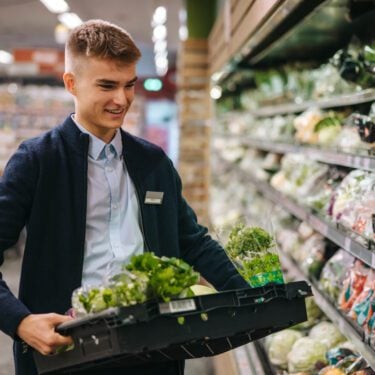 Shop assistant in supermarket re-stocking fresh vegetables in shelves of produce section.