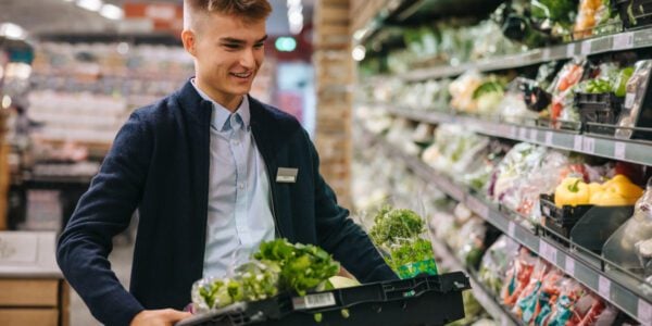 Shop assistant in supermarket re-stocking fresh vegetables in shelves of produce section.