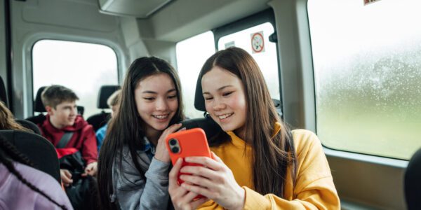A front view of a two teenage girls on the minibus on the way to go on a hiking field trip. They are looking at one of the girls mobile phones and smiling.