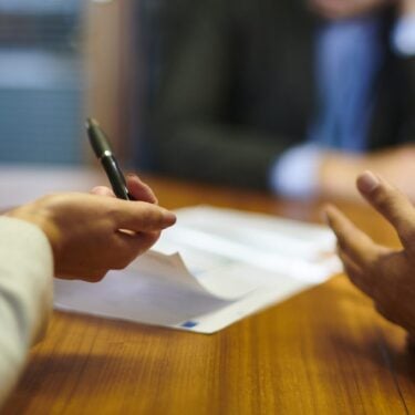 Two people have their arms on the table while they speak at a tribunal. Someone is blurred on the over side of the table.