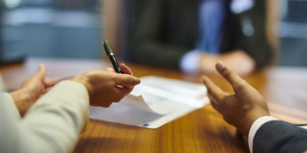 Two people have their arms on the table while they speak at a tribunal. Someone is blurred on the over side of the table.