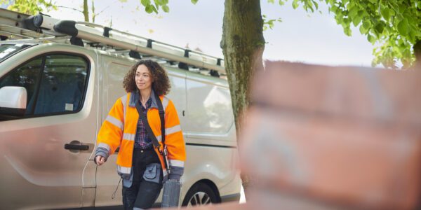 Female electrician arrives at job. She is wearing an orange hi-vis jacket and behind her is a van.