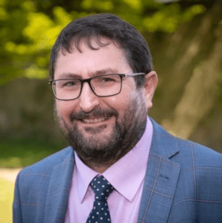 Headshot of a white man with a dark hair, a beard, and glasses. He is wearing a suit and is standing outside. He is smiling at the camera.