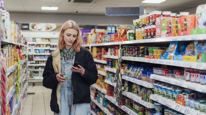 A person with light-coloured hair is standing in the aisle of a well-lit grocery store, holding a product in one hand and a smartphone in the other. They are wearing a black jacket over denim overalls. The shelves on both sides are stocked with various food items, including snacks, cereals, and canned goods.