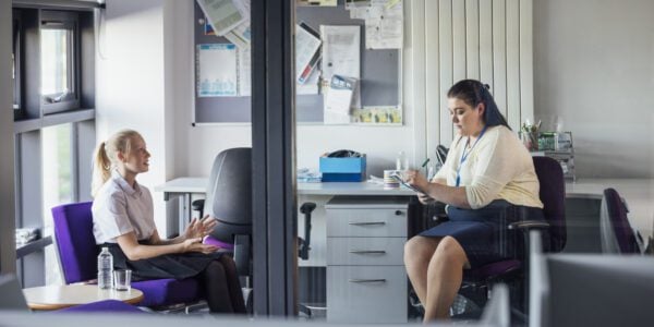A female secondary school student, sitting down in a teacher's office in a school. They are having a meeting and the teacher is taking notes while the girl talks.