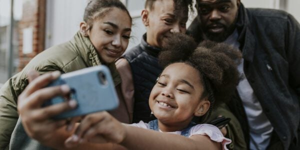 A happy young Black family poses for a selfie. The youngest daughter is at the front holding the phone.