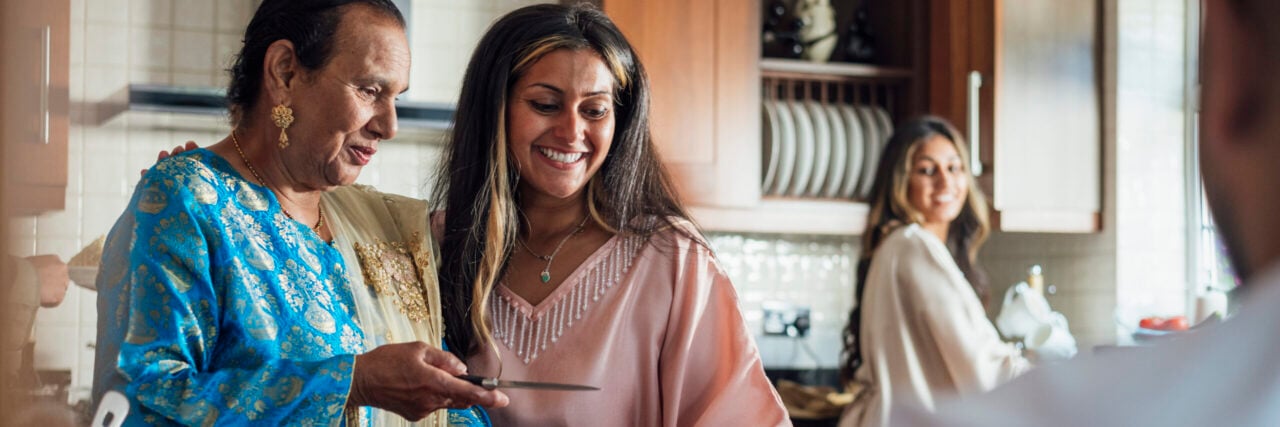 A multi-generational family gathered for Eid celebrations at home in Middlesbrough, North East England. They are all dressed in traditional outfits for the occasion. They are in the kitchen preparing food as they talk, laugh and spend quality time together.