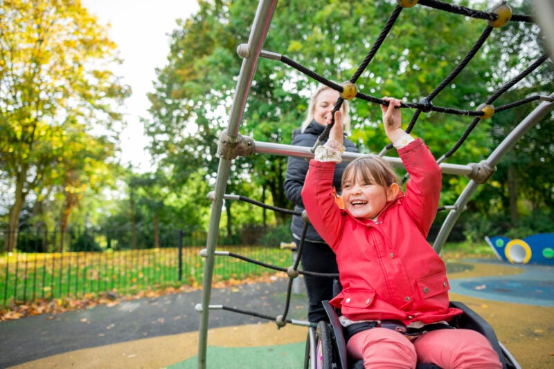 A cheerful low angle medium close-up of a young girl who is a wheelchair user playing in the park on the climbing frame with her mother. They're in a playground.