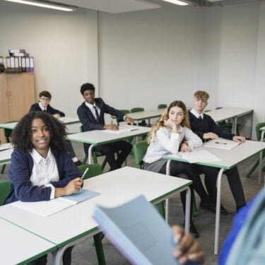Over the shoulder view of classmates pausing from writing to listen as mature Black teacher talks with teenage schoolgirl sitting in front row.