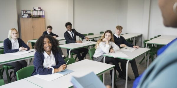 Over the shoulder view of classmates pausing from writing to listen as mature Black teacher talks with teenage schoolgirl sitting in front row.