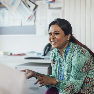 Over-the-shoulder shot of a female secondary school teacher having a one-to-one conversation with a male teenage student. They are both sitting down and she is maintaining eye contact as she is talking. The teacher is smiling and sharing a positive interaction with the student.