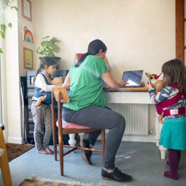 A heavily pregnant mother working at home on her laptop while talking to her kids as they stand near her.