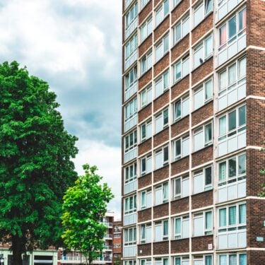 A mid-level street view of an older brick social housing block. To the left is a large green tree.