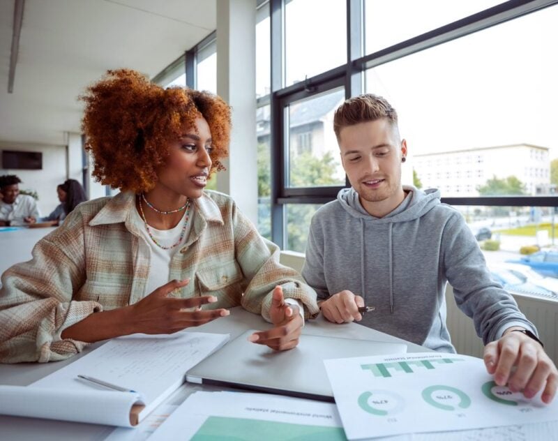 Two young adult students, one a black woman and one a white man, sit next to each other and work on a report. They are discussing and looking at papers and notes.
