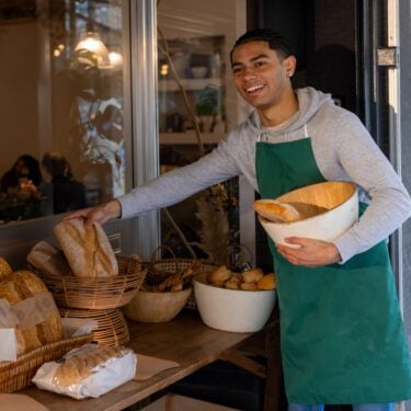 A cheerful young man wearing a green apron and a grey hoodie. He is placing fresh baked goods outside a cafe, holding a bowl filled with freshly baked bread. The cafe is in the background.