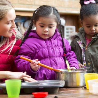 A horizontal image of two children outdoors using a big mental pan to make mud pies, while a young woman supervises. The children are both wrapped up warm in coats and are contently playing
