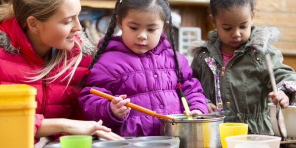 A horizontal image of two children outdoors using a big mental pan to make mud pies, while a young woman supervises. The children are both wrapped up warm in coats and are contently playing