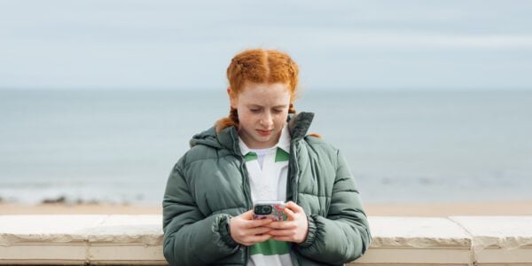 A portrait of a teenager wearing casual clothing on an overcast summer day in Whitley Bay, Northeastern England. They are standing and looking concerned as they use their smartphone.