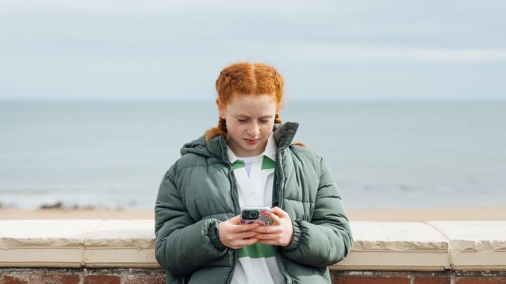 A portrait of a teenager wearing casual clothing on an overcast summer day in Whitley Bay, Northeastern England. They are standing and looking concerned as they use their smartphone.