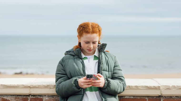 A portrait of a teenager wearing casual clothing on an overcast summer day in Whitley Bay, Northeastern England. They are standing and looking concerned as they use their smartphone.