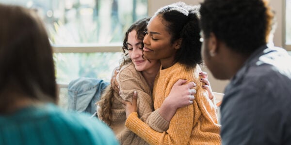 Two young women hug from the side. They are smiling and sat with a group of other young people.