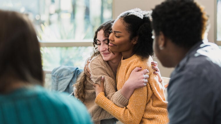 Two young women hug from the side. They are smiling and sat with a group of other young people.