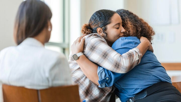 A view from behind an unrecognizable female counselor of a mother and daughter embracing and reconciling.