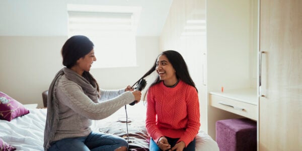 A mother and her daughter are sitting on her bed whilst she curls her daughter's hair. They both look happy to be bonding and sharing this moment together.