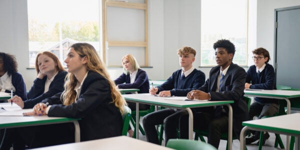 Three-quarter front view of multiracial group of students wearing uniforms, sitting side by side at desks, and listening to off-camera teacher.