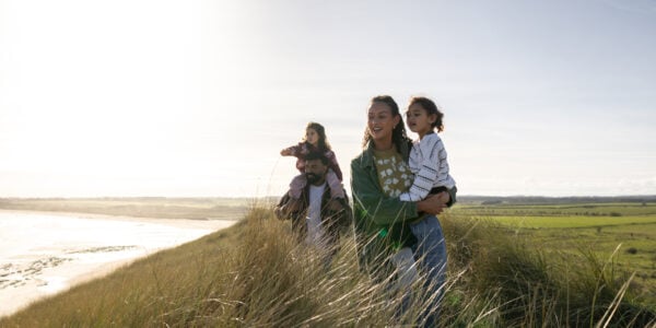 A full shot of a father, mother, and their two young daughters walking together through tall dunes near the coast. Both of the daughters are being carried. The low, warm sunlight lights up the whole scene, creating a peaceful, tranquil atmosphere
