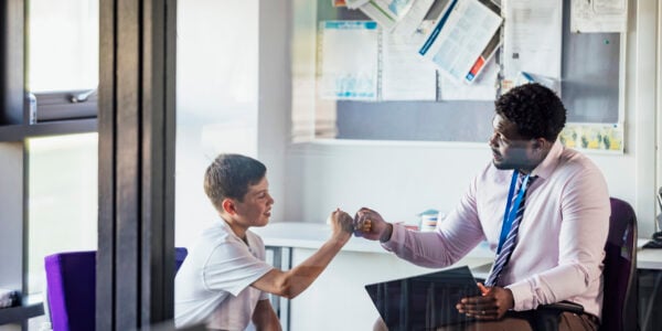 A male secondary school student sitting in an office in the school, having a meeting with his teacher. They are giving each other a fist bump after a successful meeting.